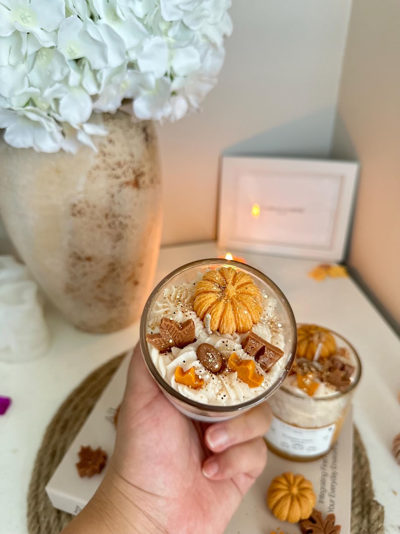 Puede incluir: Un recipiente de vidrio para velas relleno de cera blanca con una calabaza naranja decorativa, hojas de arce marrones y peque&ntilde;as incrustaciones de cera en forma de estrella. La superficie tiene purpurina dorada y textura batida.