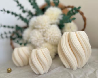 Puede incluir: Tres jarrones de cerámica color crema, con remolinos, de diferentes tamaños, están dispuestos sobre una superficie clara. Un cesto lleno de flores de pompón blancas y ramitas verdes está en el fondo. Dos pequeñas cuentas decorativas doradas están dispersas.