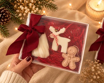 Puede incluir: Una caja de regalo blanca con tapa transparente y lazo rojo oscuro que contiene tres figuras de cera: un árbol de Navidad blanco, un reno blanco con cordel rojo y blanco, y un hombre de jengibre marrón claro sobre virutas de papel rojo.