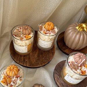 Puede incluir: Tres velas en frasco de vidrio con cera blanca, decoradas con peque&ntilde;as figuras de cera en forma de calabaza naranja y marr&oacute;n y hojas de oto&ntilde;o. Cada etiqueta dice La Belle Lumiere Pumpkin Spice. Tambi&eacute;n hay dos velas s&oacute;lidas con forma de calabaza.
