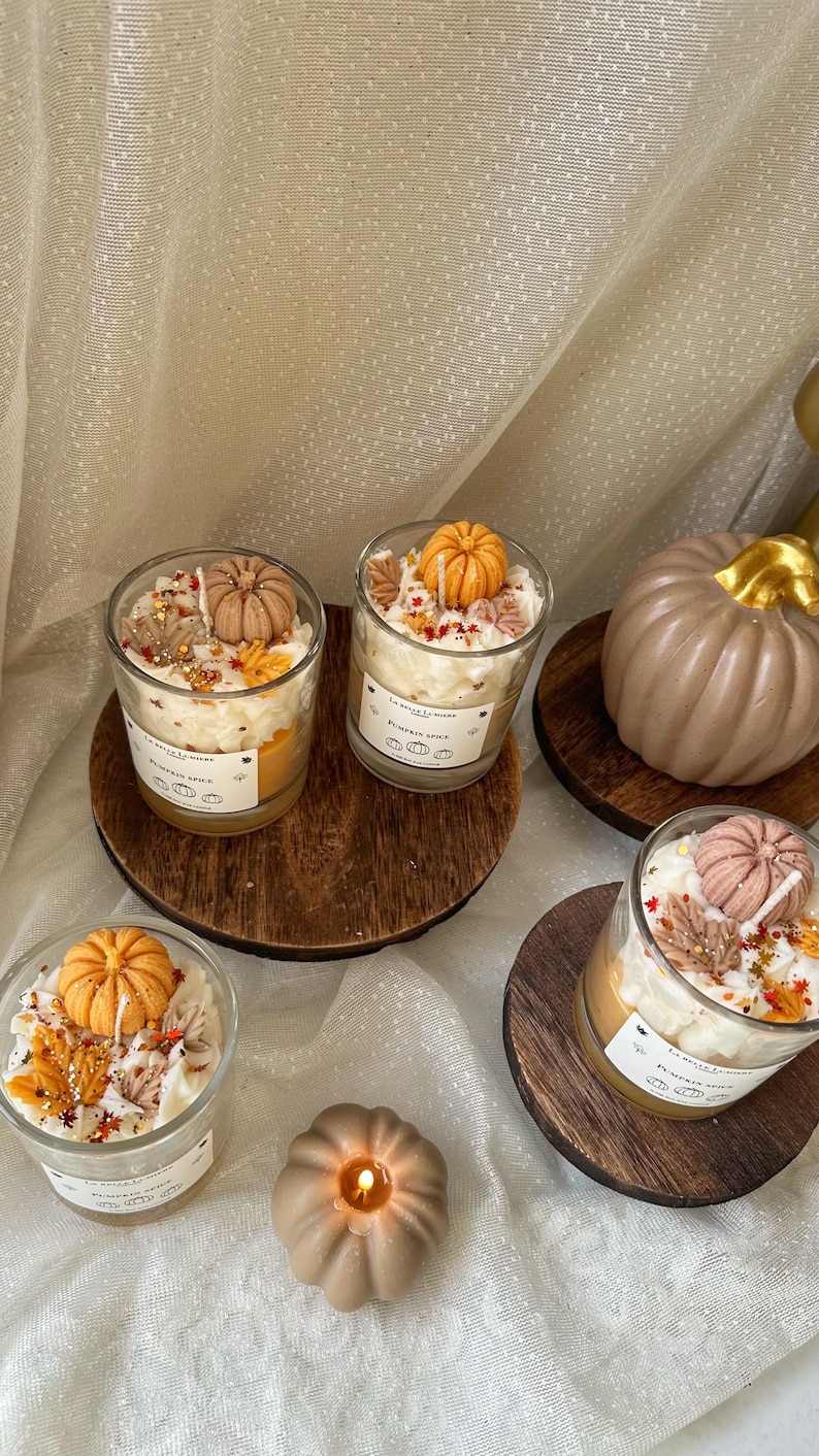 Puede incluir: Tres velas en frasco de vidrio con cera blanca, decoradas con peque&ntilde;as figuras de cera en forma de calabaza naranja y marr&oacute;n y hojas de oto&ntilde;o. Cada etiqueta dice La Belle Lumiere Pumpkin Spice. Tambi&eacute;n hay dos velas s&oacute;lidas con forma de calabaza.