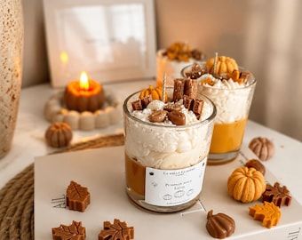 Puede incluir: Dos velas de cristal con cimas de cera batida blanca, decoradas con pequeñas calabazas de cera y piezas color chocolate. La etiqueta frontal dice La Belle Lumiere, Pumpkin Spice Latte. Hojas y calabazas de cera dispersas rodean las velas.