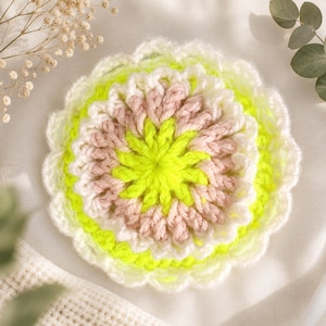 Peut inclure: Un dessous de verre circulaire au crochet présentant un motif floral superposé. Le centre est d'un jaune néon vibrant, entouré d'un anneau de fil rose poudré doux, suivi d'une bordure extérieure en fil blanc avec un bord festonné.