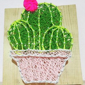 Peut inclure: Une œuvre d'art en fil représentant un cactus sur une planche de bois. Le cactus est réalisé avec du fil vert et jaune, surmonté d'une fleur rose. Il est placé dans un pot rose, également en fil, avec un contour blanc.