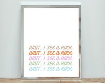 Op de afbeelding: Een ingelijst afdruk met de tekst "WAIT, I SEE A ROCK" die vijf keer wordt herhaald in verschillende pastelkleuren. De tekst is in een retro-lettertype stijl.