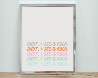 Op de afbeelding: Een ingelijst print met de tekst "WAIT, I SEE A ROCK" die vijf keer wordt herhaald in verschillende pastelkleuren. De tekst is in een retro-lettertype stijl.