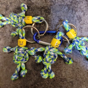 May include: Four keychains with blue and green paracord figures attached to silver key rings. Each figure has a yellow face with a different expression.