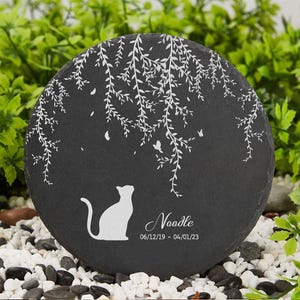 May include: A round, black slate stone with a white cat silhouette and the name "Noodle" and dates 06/12/19 - 04/01/23. The stone features a white tree branch design and butterflies.