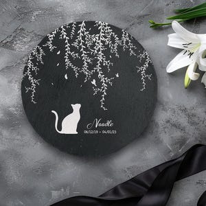 May include: A round, black memorial stone with a white cat silhouette and the name "Noodle" and dates. The stone features a white weeping willow design and butterflies. A white lily and black ribbon are in the background.