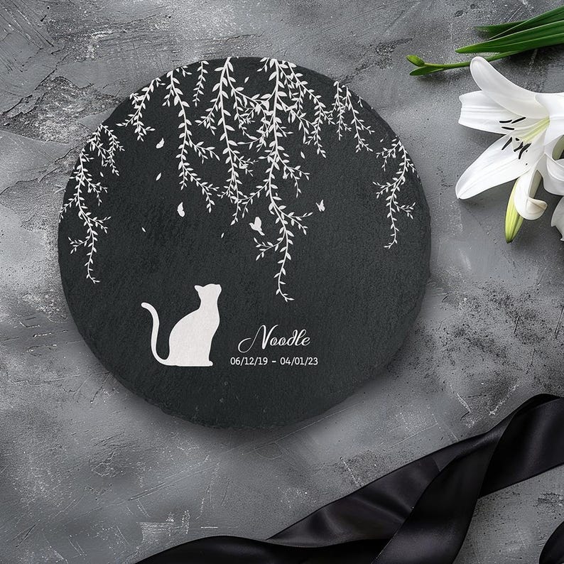 May include: A round, black memorial stone with a white cat silhouette and the name "Noodle" and dates. The stone features a white weeping willow design and butterflies. A white lily and black ribbon are in the background.