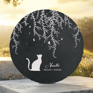 May include: A round, black memorial stone with a white silhouette of a cat looking up. White branches and butterflies are etched around the top. The name "Noodle" and dates 06/12/19 - 04/01/23 are below the cat.