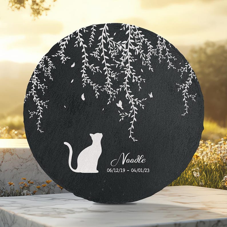 May include: A round, black memorial stone with a white silhouette of a cat looking up. White branches and butterflies are etched around the top. The name "Noodle" and dates 06/12/19 - 04/01/23 are below the cat.
