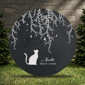 May include: A round, black memorial stone with a white cat silhouette looking up. White weeping willow branches and butterflies are etched above the cat. The name "Noodle" and dates 06/12/19 - 04/01/23 are below the cat.