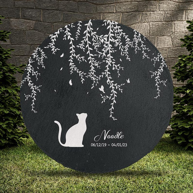 May include: A round, black memorial stone with a white cat silhouette looking up. White weeping willow branches and butterflies are etched above the cat. The name "Noodle" and dates 06/12/19 - 04/01/23 are below the cat.