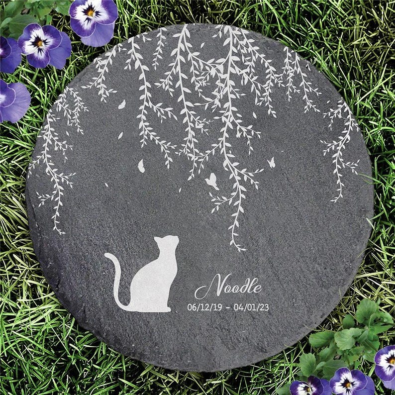 May include: A round, dark gray stone memorial with a white cat silhouette, weeping willow design, and the name "Noodle" with dates. The stone is set in green grass with purple flowers.