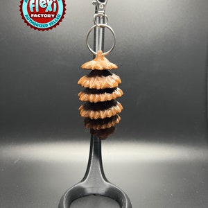 May include: A brown, 3D printed keychain shaped like a pine cone with a silver metal key ring attached.