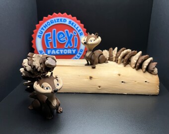 May include: Two brown and tan squirrel figurines with bushy tails. The squirrel on the left is holding a brown and tan pine cone. The squirrel on the right is standing on a brown log with a brown and tan tail behind it. The log is on a black surface. A red, white, and blue sticker with the text "Authorized Seller Flexi Factory" is behind the squirrels.