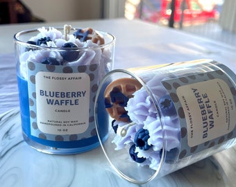 May include: Two glass candle jars with blue and purple wax, topped with blueberries and a waffle. The labels read "The Flossy Affair Blueberry Waffle Candle Natural Soy Wax Handpoured in California 10 oz."