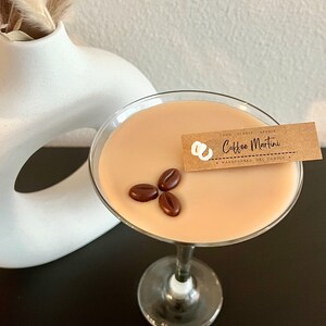 May include: A brown glass martini glass filled with a light brown scented candle. The candle is topped with three chocolate coffee beans and has a brown paper tag that reads "The Flopsy Affair Coffee Martini Handpoured Gel Candles".