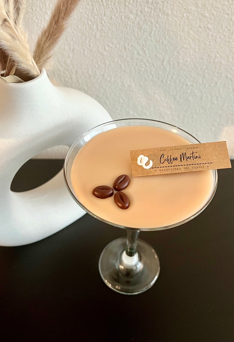 May include: A brown glass martini glass filled with a light brown scented candle. The candle is topped with three chocolate coffee beans and has a brown paper tag that reads "The Flopsy Affair Coffee Martini Handpoured Gel Candles".