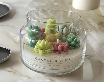 以下が含まれることがあります： 白いラベルに「The Flossy Affair Home Collection Cactus & Jade Soy Wax Candle 7oz/200g 30 Hours」と書かれた透明なガラス製のキャンドル。キャンドルは、緑、黄色、ピンクの小さなワックス製の多肉植物でいっぱいになっています。