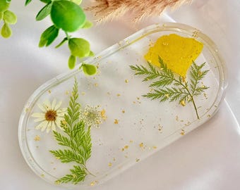 May include: Clear resin tray with embedded dried flowers and gold flakes. The tray features a yellow flower, green fern-like leaves, and a white daisy. The oval-shaped tray is a decorative item.