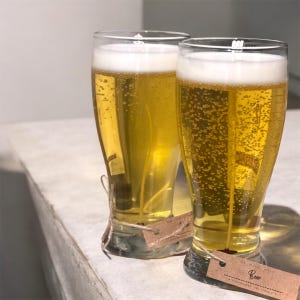 May include: Two beer-shaped candles with a foamy top and a golden-yellow body. Each candle is in a clear glass and has a small brown tag tied around the base. The tag reads "Beer".