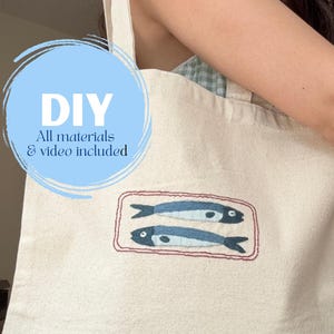 May include: Cream-colored tote bag with embroidered blue fish design. The bag has a rectangular design with a red border around the fish. The text "DIY All materials & video included" is in a blue circle.
