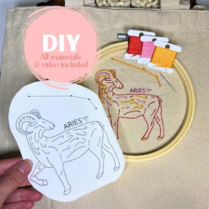 May include: DIY embroidery kit featuring an Aries zodiac design. The kit includes a beige fabric with an embroidered ram, a wooden hoop, colorful thread spools, and a printed pattern. The text "DIY All materials & video included" is in a pink circle.