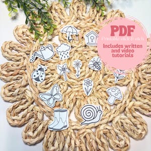 Op de afbeelding: Een verzameling zwart-witte stickers met lentethema, waaronder een gieter, lieveheersbeestje, vlinder en paddenstoel, gerangschikt op een geweven placemat. Een roze cirkel met de tekst "PDF Printable stick & stitch" staat in de afbeelding. Inclusief geschreven en video tutorials.