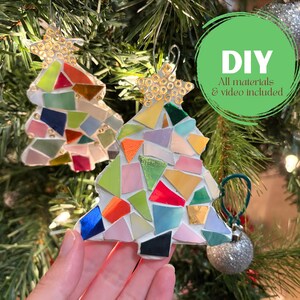 May include: Two mosaic Christmas tree ornaments with star toppers. The trees are made of colorful, irregularly shaped tiles in shades of red, green, blue, and gold. The ornaments hang on a Christmas tree with a silver ornament.