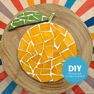 May include: A mosaic orange fruit with a green leaf, set on a wooden plate. The orange is made of yellow and orange tiles with white grout. A blue circle with the text "DIY All materials & video included" is on the bottom right.