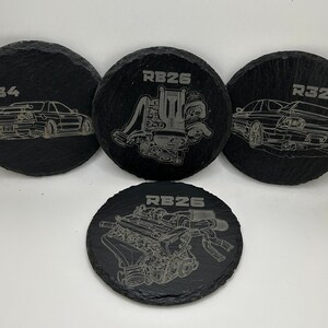 May include: Four black slate coasters with white engraved designs. The coasters feature the text "RB26", "R32", "R34", and a stylized image of an engine.