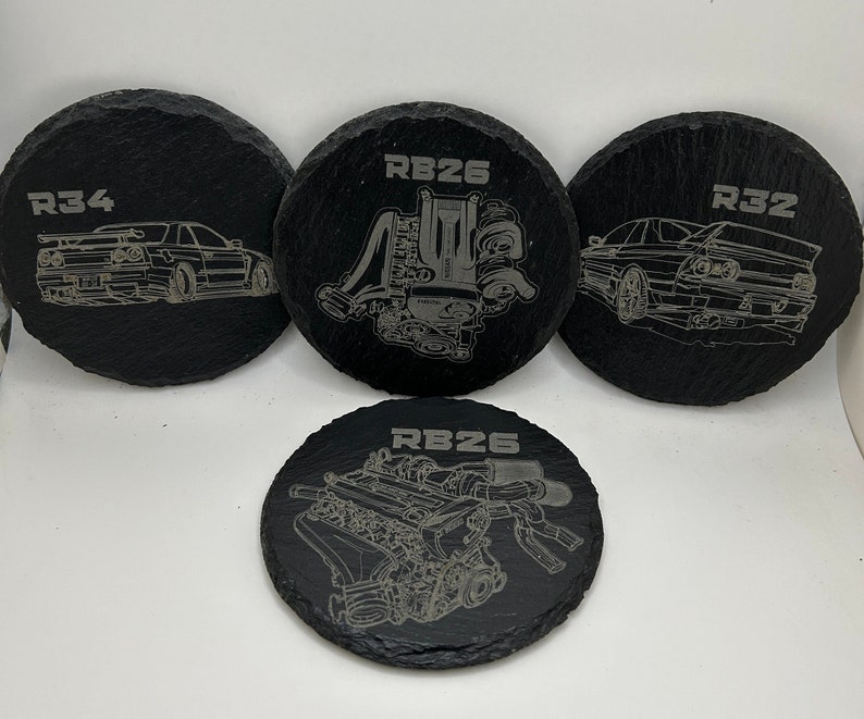 May include: Four black slate coasters with white engraved designs. The coasters feature the text "RB26", "R32", "R34", and a stylized image of an engine.
