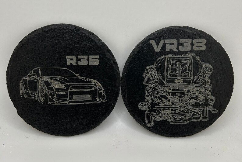 May include: Two black round slate coasters with white engraved designs. One coaster features a car with the text "R35" and the other coaster features an engine with the text "VR38".