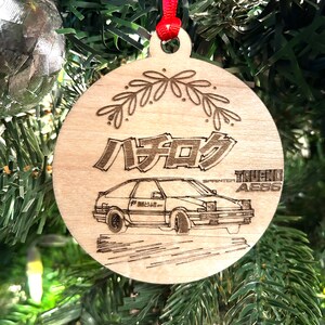 May include: A wooden Christmas ornament featuring a black and white illustration of a Toyota Sprinter Trueno AE86 car with the Japanese text "ハチロク" and the words "SPRINTER TRUENO AE86".