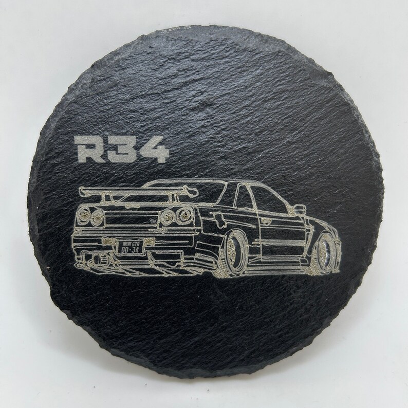 May include: A black slate coaster with a white outline of a Nissan Skyline R34 car and the text "R34" on the top.