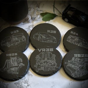 May include: Six black slate coasters with different engraved designs. The coasters feature car models and engine diagrams with the text "R32", "R35", "R34", "RB26", "VR38", and "RB26".