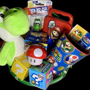 Pu&ograve; includere: Un peluche verde Yoshi, una scatola per il pranzo rossa con un personaggio di Super Mario, una tazza blu con un personaggio di Super Mario, un giocattolo a forma di fungo rosso e bianco e vari dolci e leccornie a tema Super Mario in un cestino verde.
