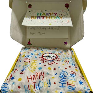 May include: A yellow gift box with a white tissue paper lining. The tissue paper has a colorful pattern of the words "Happy Birthday" and stars. The box is open and a card is visible inside. The card says "Happy Birthday Charlie!! From: Manu".