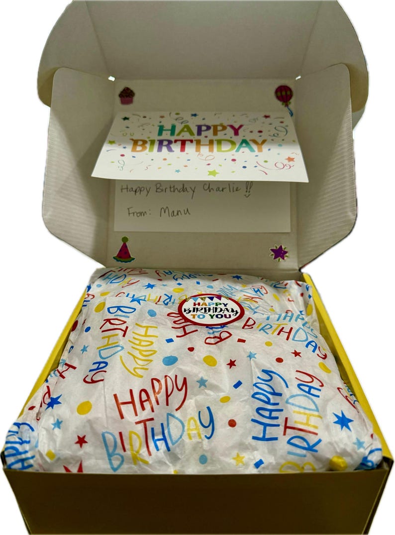 May include: A yellow gift box with a white tissue paper lining. The tissue paper has a colorful pattern of the words "Happy Birthday" and stars. The box is open and a card is visible inside. The card says "Happy Birthday Charlie!! From: Manu".