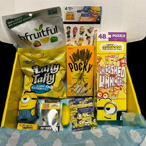May include: A yellow gift box filled with Minion themed snacks and toys. The box contains a bag of fruit snacks, a bag of Laffy Taffy candy, a box of Pocky sticks, a Minion puzzle, a Minion sticker sheet, a Minion plush toy, a banana-shaped gummy candy, and a Minion keychain.