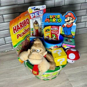 Pu&ograve; includere: Un cesto verde e bianco pieno di regali a tema Super Mario e Donkey Kong. Il cesto include un Donkey Kong di peluche, una tazza di Super Mario, un dispenser di caramelle di Super Mario, una confezione di gommose alla pesca Haribo e un blocco interrogativo di Super Mario.