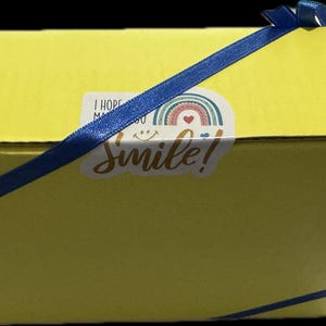 May include: A yellow cardboard box with a blue ribbon tied around it. The box has a sticker on the top that says "I hope you smile!" with a rainbow and a heart.