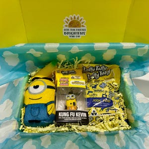 May include: A yellow gift box with a blue and white patterned lining. The box contains a yellow plush Minion toy, a yellow Laffy Taffy candy bag, a yellow Minion keychain, and a yellow and blue banana-shaped plush toy. The box has a sticker on the top that says "Hope this package brightens your day!"