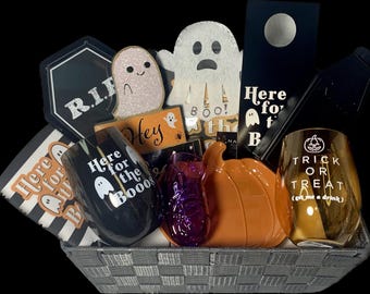 May include: A Halloween-themed gift basket filled with decorative items. Includes two wine glasses with ghost and pumpkin designs, a ceramic pumpkin, and various signs with phrases like "Here for the Booos" and "Trick or Treat."