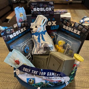 May include: A blue basket filled with Roblox-themed gifts. Includes a foil-wrapped chocolate bunny, Roblox game cards, and toy figures. The basket also contains candy and a box with the text "ONLY THE TOP GAMES!"