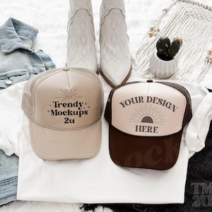 May include: Two trucker hats, one brown and one beige, with white mesh backs. The beige hat has the text 'Trendy + Mockups 2u' on it. The brown hat has the text 'Your Design Here' on it. The hats are laying on a white surface with a white t-shirt, a blue denim jacket, and a pair of black denim shorts.