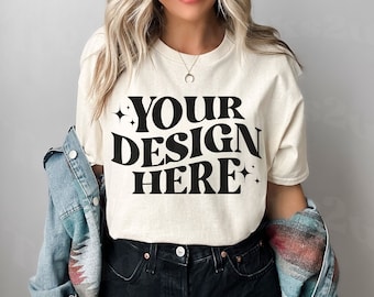 May include: A woman wearing a light beige t-shirt with the text "YOUR DESIGN HERE" in black. She is also wearing black denim shorts and a blue denim jacket.