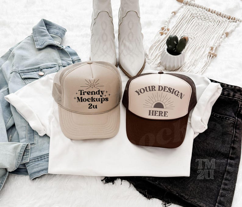 May include: Two trucker hats, one brown and one beige, with white mesh backs. The beige hat has the text 'Trendy + Mockups 2u' on it. The brown hat has the text 'Your Design Here' on it. The hats are laying on a white surface with a white t-shirt, a blue denim jacket, and a pair of black denim shorts.